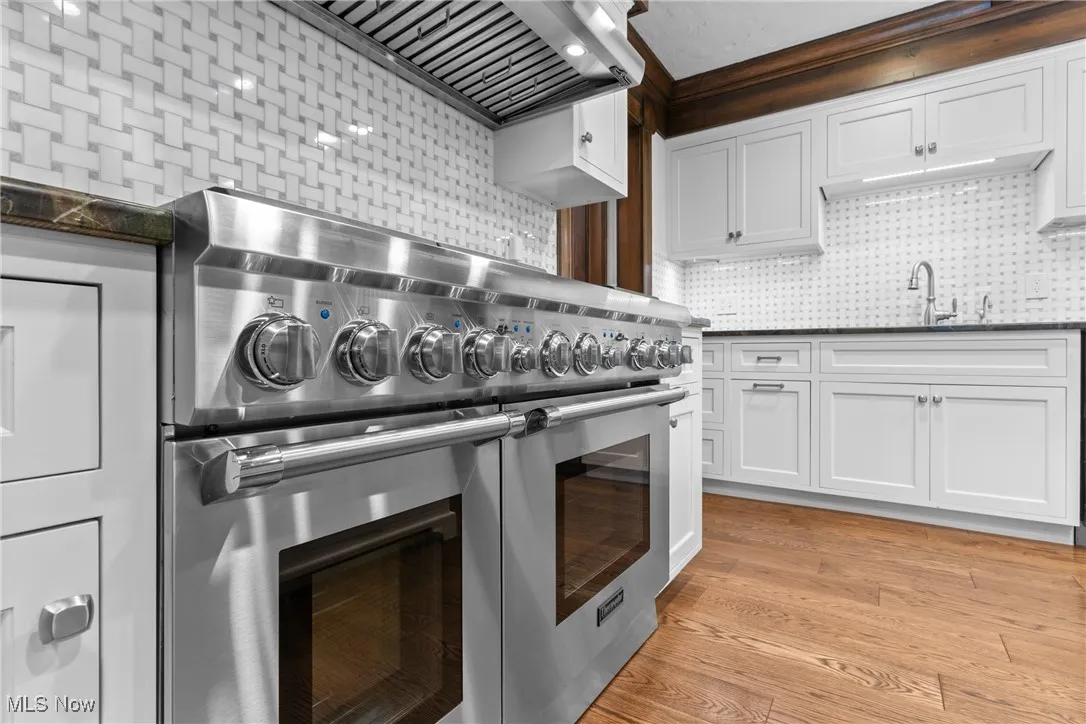 Kitchen with tasteful backsplash, wall chimney exhaust hood, range with two ovens, light wood-style floors, and white cabinetry