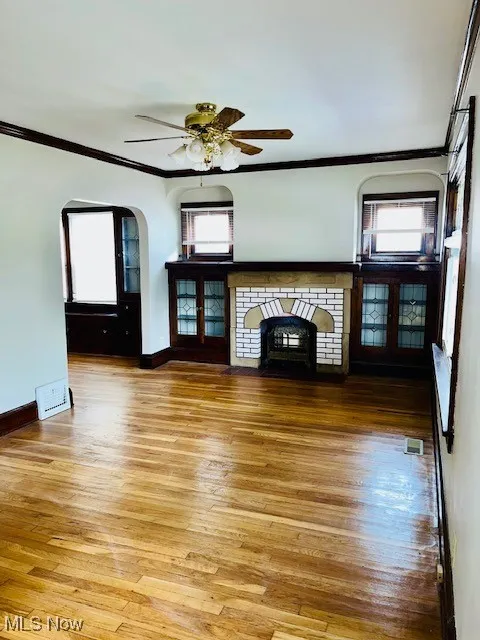 Unfurnished living room with arched walkways, crown molding, light wood finished floors, a ceiling fan, and a fireplace