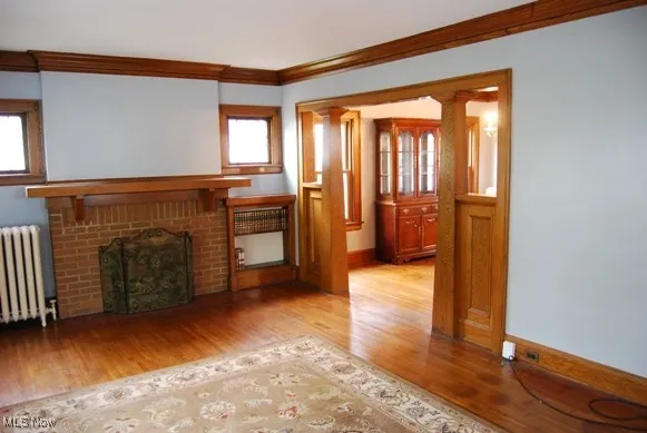 Unfurnished living room featuring radiator heating unit, crown molding, light wood finished floors, a brick fireplace, and ornate columns