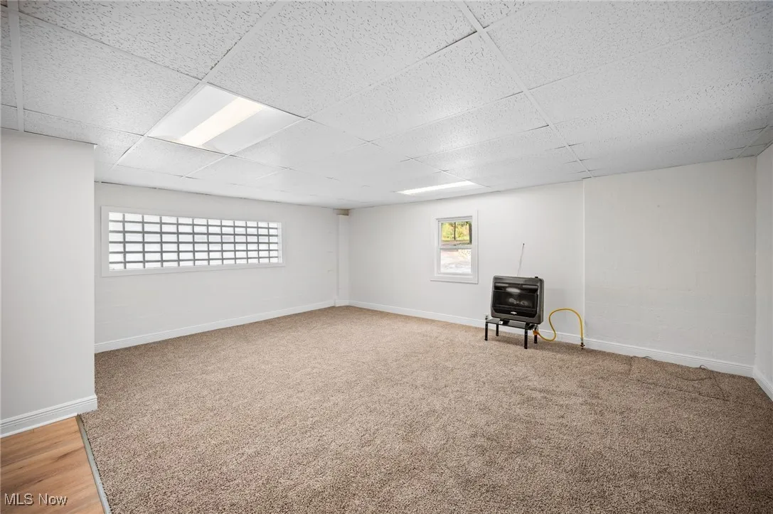 Basement featuring heating unit, light colored carpet, a paneled ceiling, and a wood stove