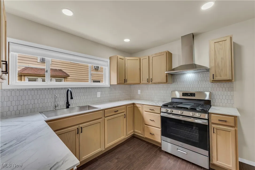 Kitchen with stainless steel gas stove, wall chimney exhaust hood, light brown cabinetry, decorative backsplash, and dark wood-style flooring