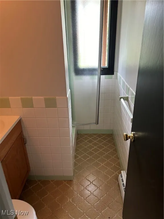 Bathroom with tile walls, vanity, wainscoting, a shower stall, and a baseboard radiator