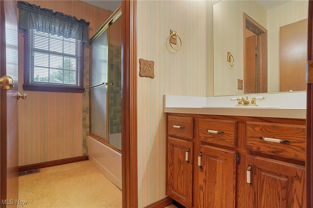 Full bathroom featuring bath / shower combo with glass door, wallpapered walls, and vanity