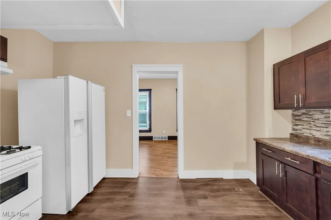 Kitchen with dark brown cabinets, dark wood-style flooring, white appliances, backsplash, and dark countertops