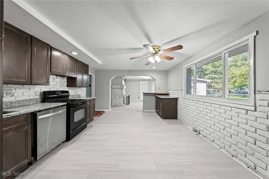 Kitchen featuring dark brown cabinets, brick wall, black gas range, stainless steel dishwasher, and light stone counters
