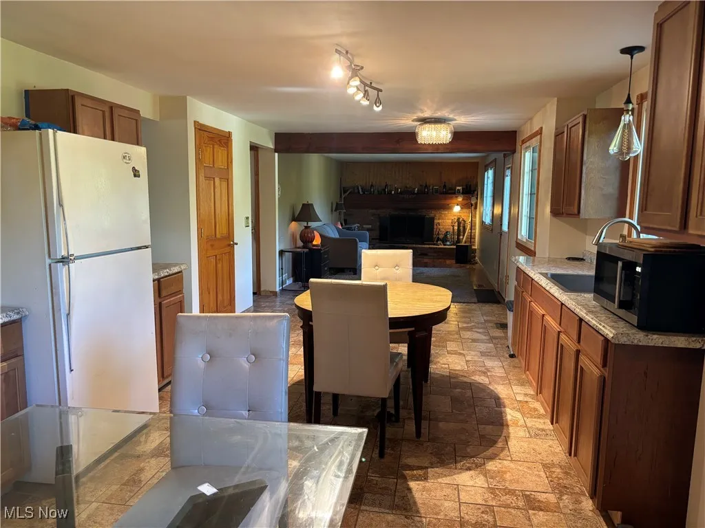 Kitchen with freestanding refrigerator, brown cabinetry, black microwave, pendant lighting, and stone tile floors