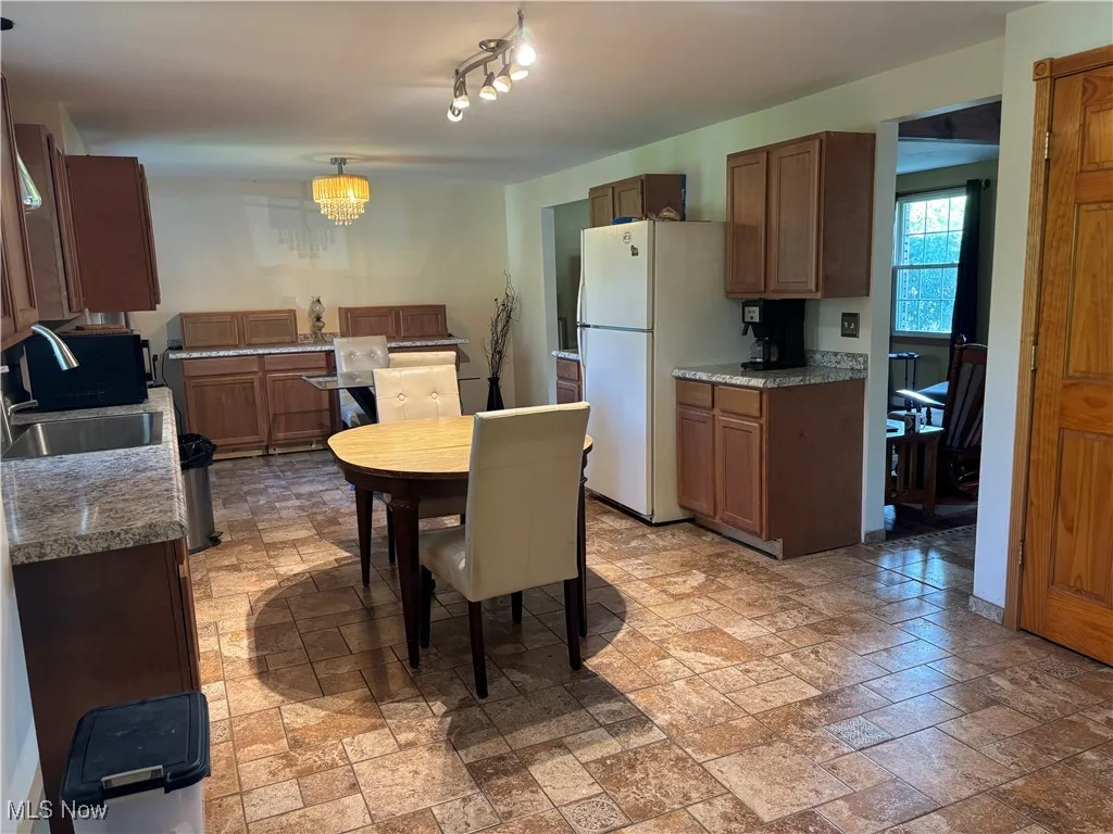 Kitchen featuring freestanding refrigerator, brown cabinetry, light stone finish flooring, hanging light fixtures, and a chandelier