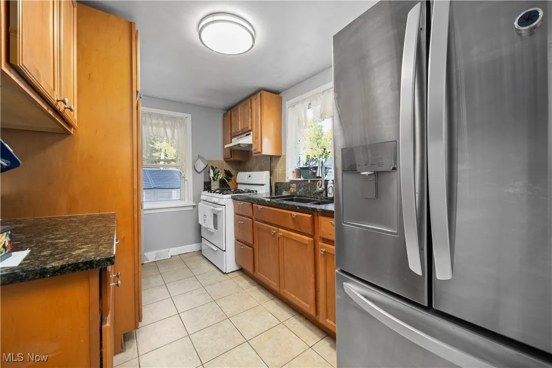 Kitchen featuring stainless steel fridge, white gas range, light tile patterned flooring, brown cabinets, and dark stone countertops