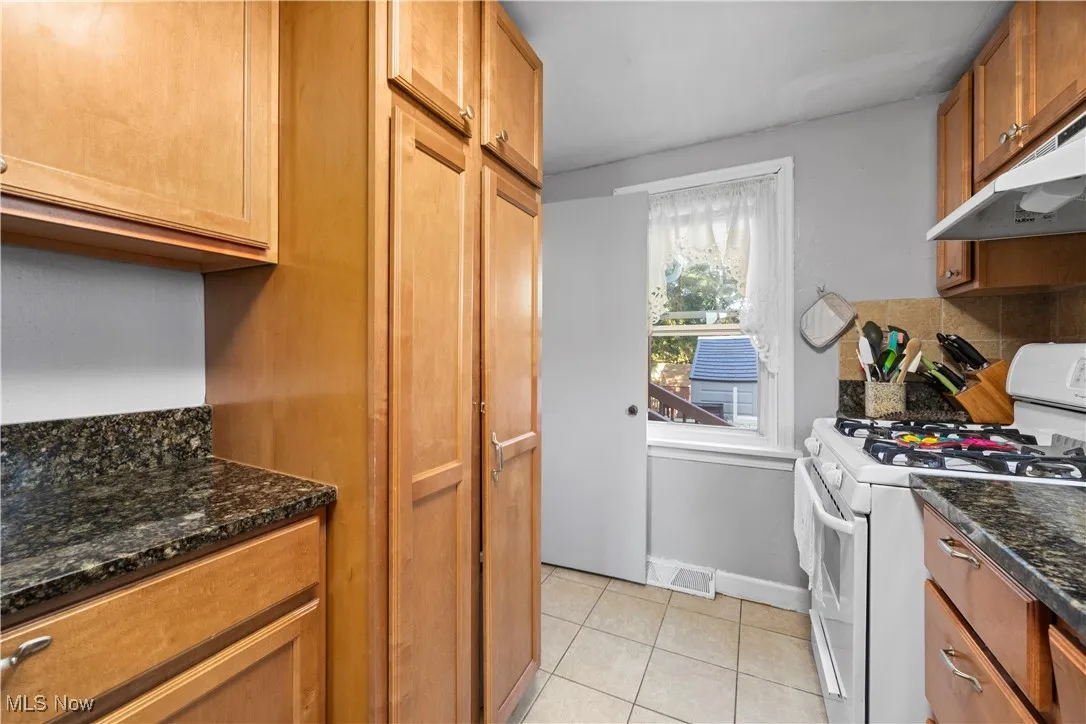 Kitchen featuring white gas range, light tile patterned flooring, dark stone counters, brown cabinetry, and decorative backsplash