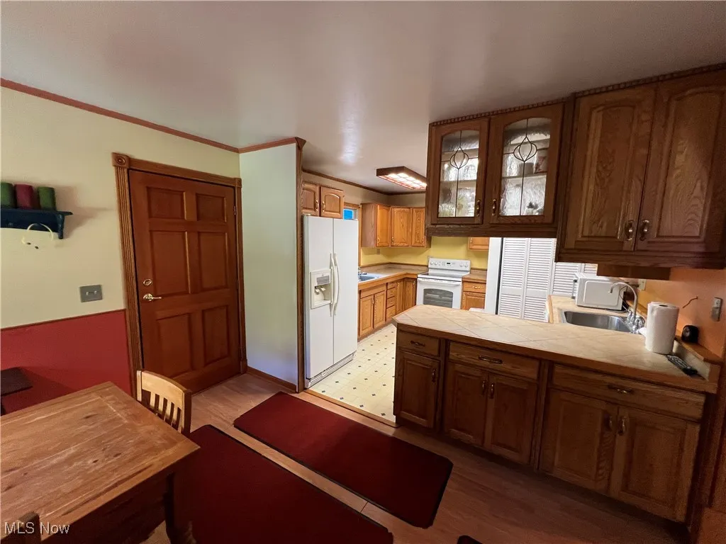 Kitchen with glass insert cabinets, white appliances, crown molding, light wood-style floors, and tile counters
