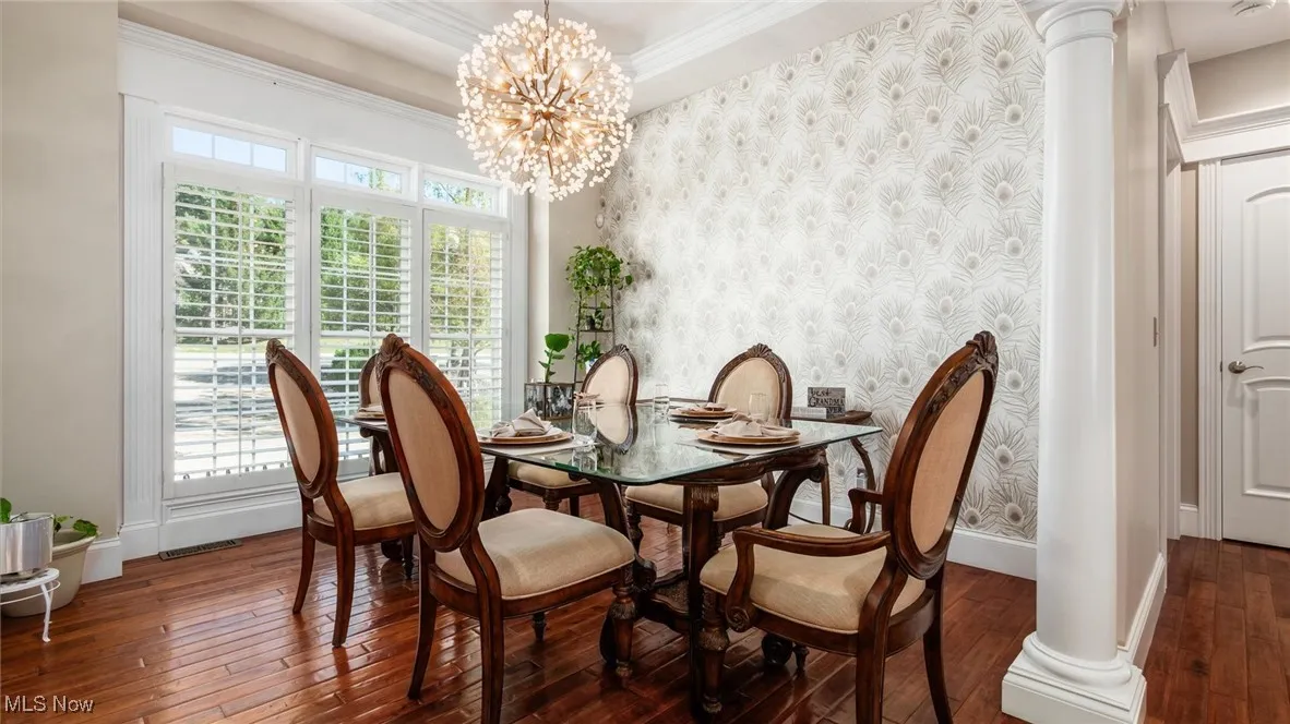 Dining area featuring wallpapered walls, dark wood-style floors, a chandelier, decorative columns, and ornamental molding