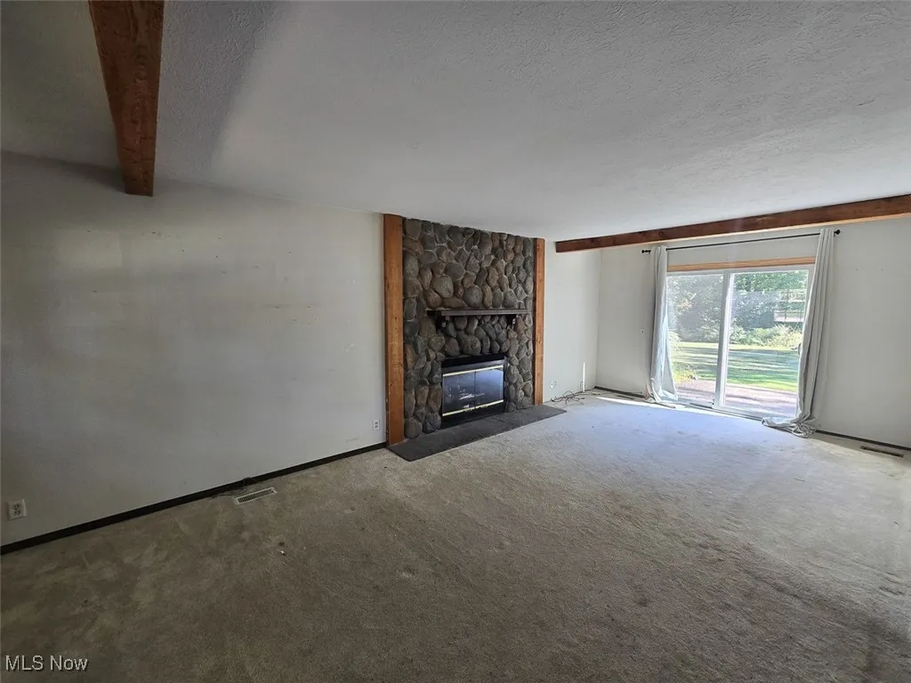 Unfurnished living room featuring a textured ceiling, a stone fireplace, carpet floors, and beam ceiling