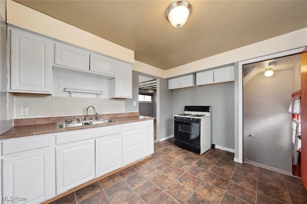 Kitchen featuring white range with gas cooktop, backsplash, stone finish flooring, and white cabinetry