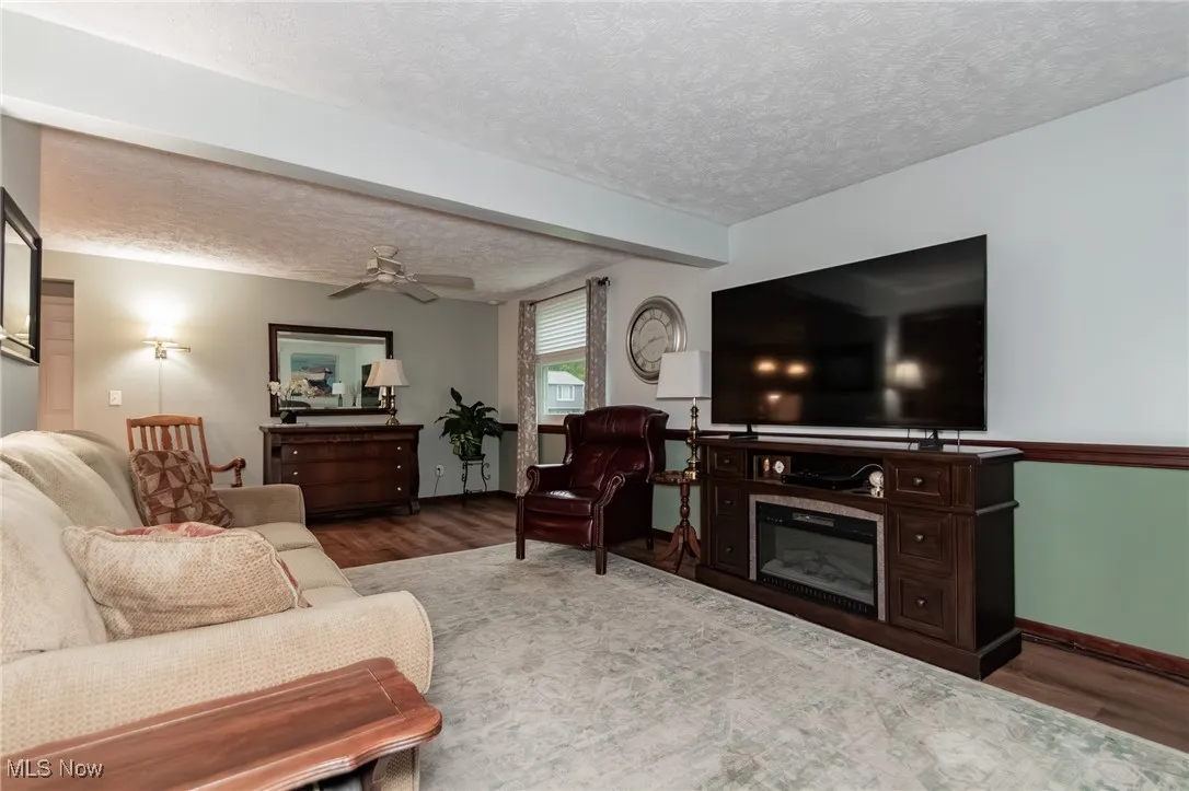 Living area featuring a textured ceiling, a ceiling fan, beamed ceiling, wood finished floors, and a glass covered fireplace