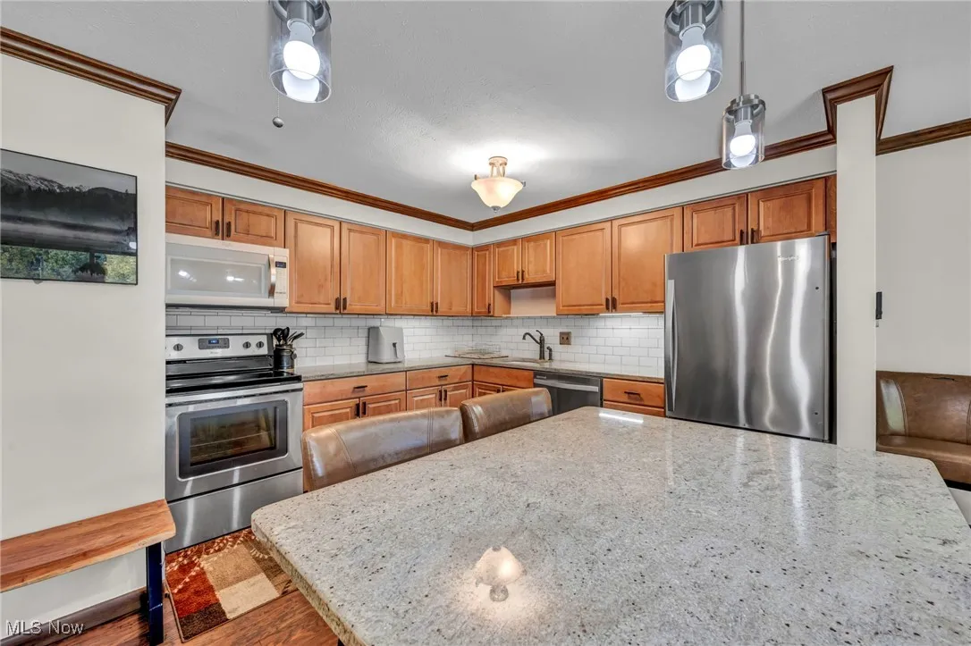 Kitchen with stainless steel appliances, decorative backsplash, crown molding, light stone countertops, and brown cabinetry