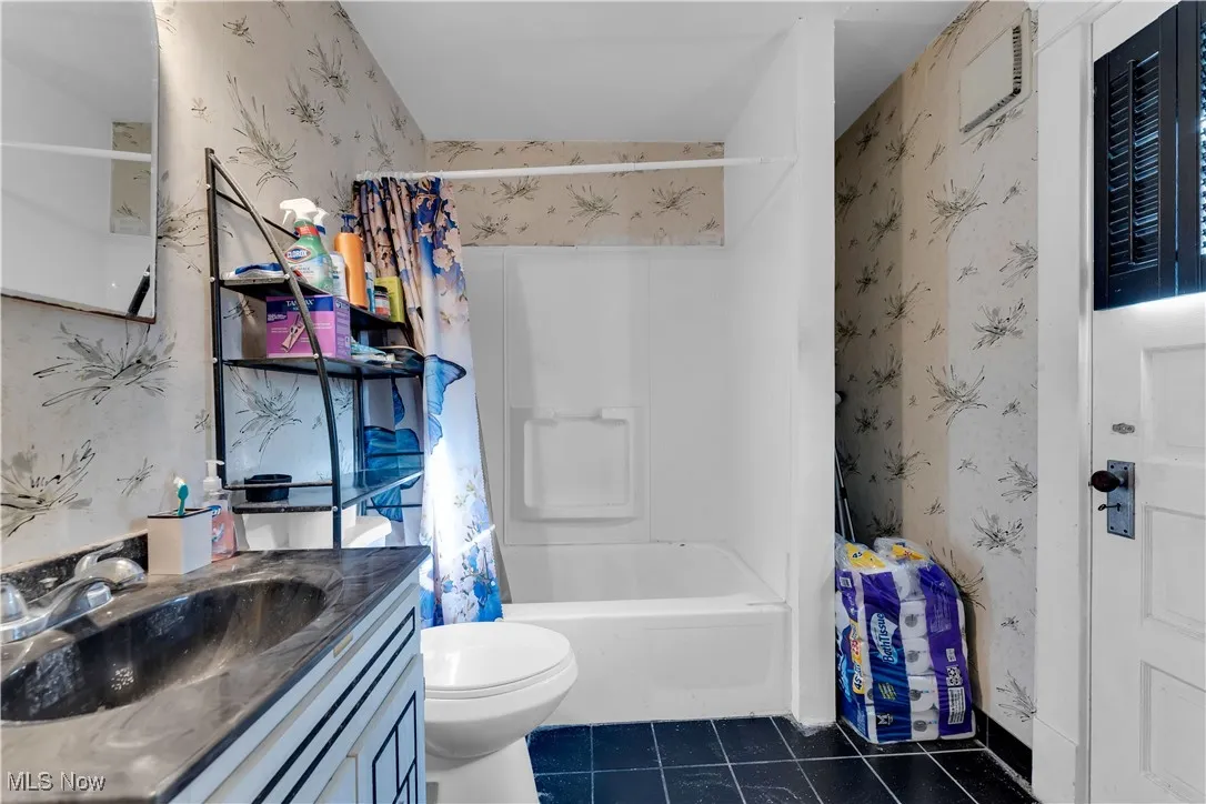 Full bathroom with wallpapered walls, dark tile patterned floors, vanity, and shower / bath combination with curtain