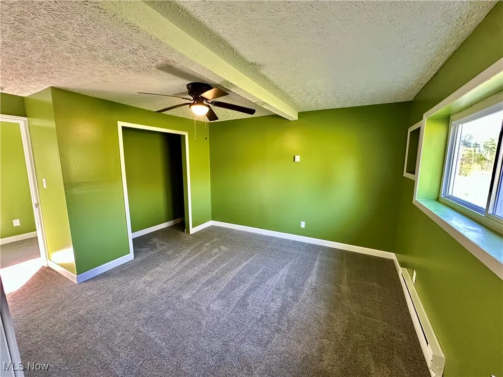 Unfurnished bedroom featuring dark carpet, a textured ceiling, a baseboard heating unit, a closet, and a ceiling fan