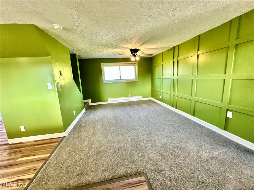 Carpeted empty room featuring a textured ceiling, ceiling fan, a baseboard radiator, a decorative wall, and a smoke detector