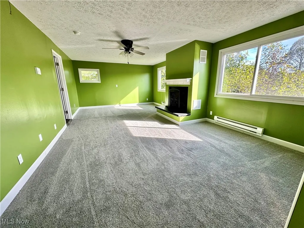 Unfurnished living room with carpet, baseboard heating, a ceiling fan, a textured ceiling, and a fireplace with raised hearth