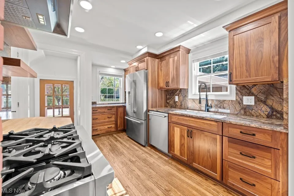 In this sunlit culinary dream, the walnut cabinets stand tall like wizards of warmth, their rich grain whispering secrets to the room. A gooseneck faucet twirls above a stainless steel sink that gleams like a polished moon in a twilight puddle. The warm brown marble on the backsplash and countertops rolls with storm-cloud veins of caramel and espresso, as if a friendly dragon drew breezy map lines across the surface.

The kitchen wears a ribbon of stainless steel appliances, marching in neat parade formation, ready for a grand feast or a quiet tea party for one. Light spills from the big window, dancing across the hardwood floors and turning every utensil handle into a tiny sunbeam.

With the cabinetry as a sturdy chorus and the marble as the melodious chorus line, this kitchen invites you to cook, dream, and maybe hum a tune to the rhythm of sizzling pans and simmering sauces.