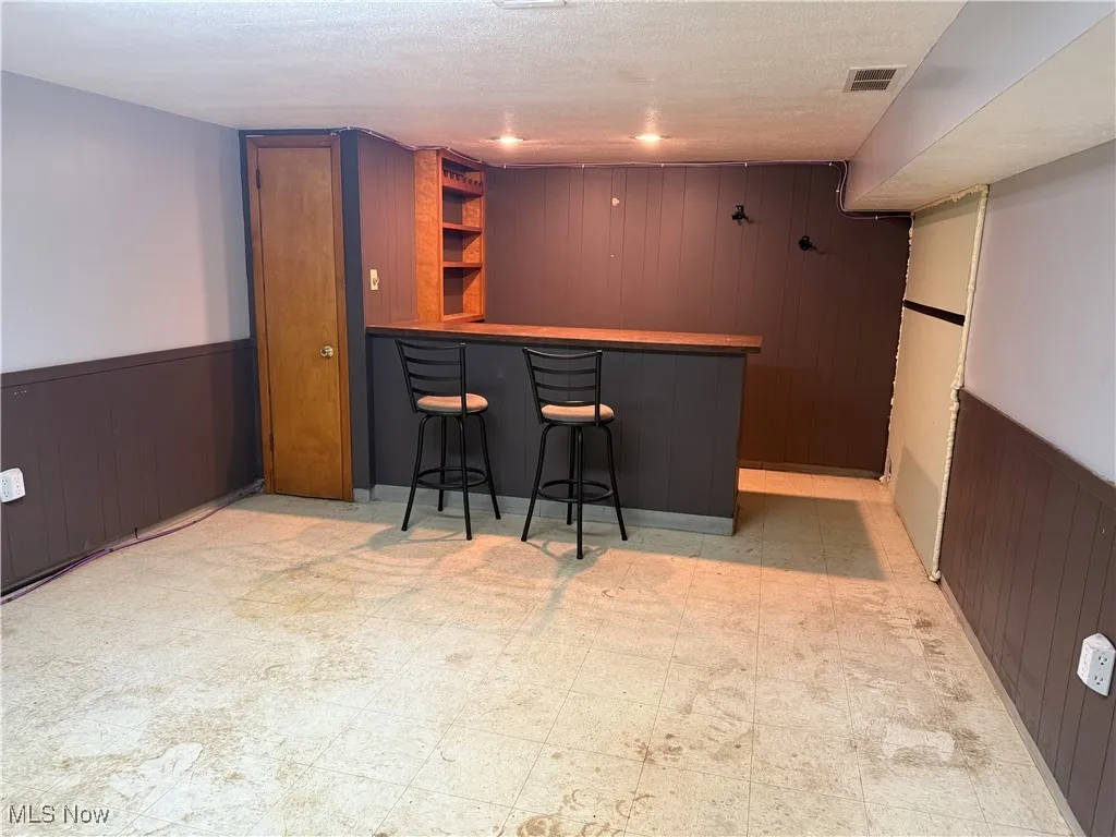 Basement - Indoor dry bar with wood walls, a wainscoted wall, open shelves, light floors, and a textured ceiling