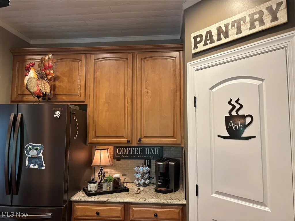 Kitchen with freestanding refrigerator, tasteful backsplash, light stone countertops, brown cabinetry, and crown molding