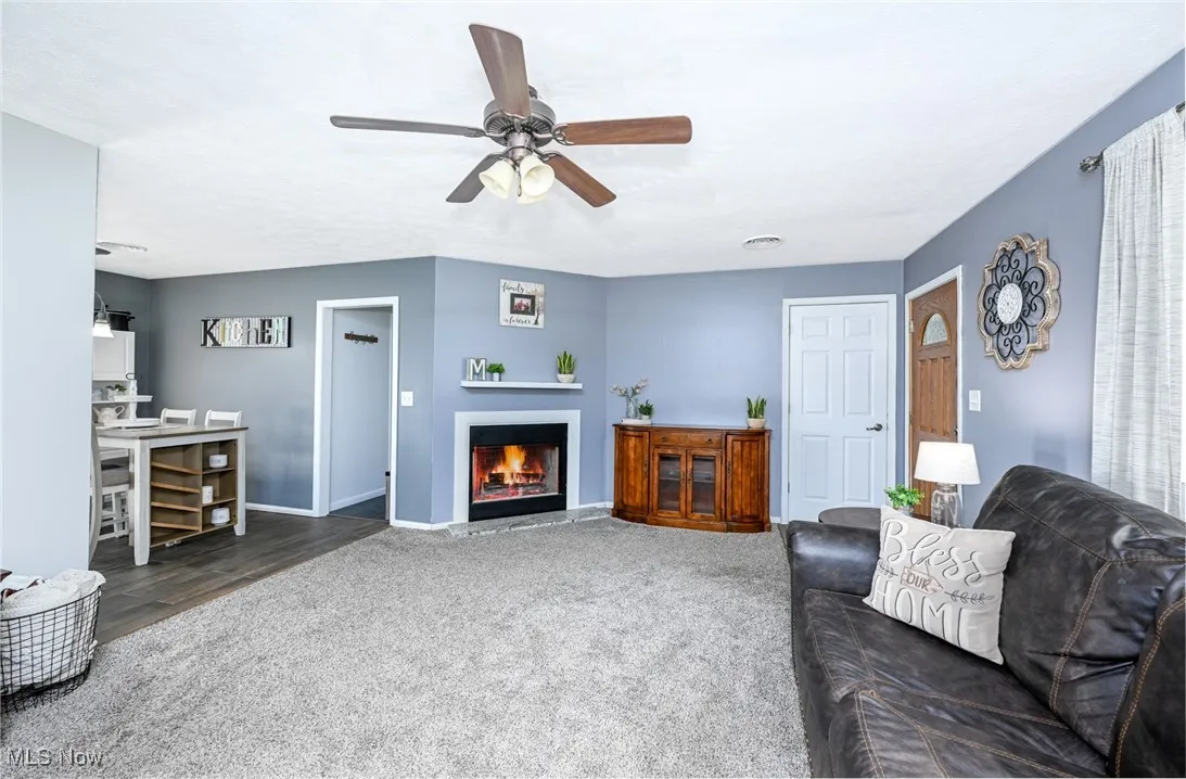 Living area featuring carpet, a warm lit fireplace, and a ceiling fan