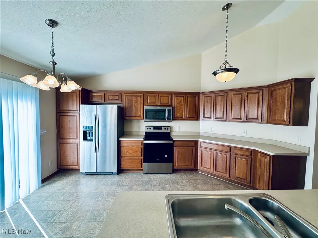 Kitchen with stainless steel appliances, brown cabinetry, stone finish flooring, light countertops, and a chandelier.