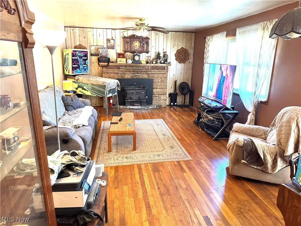 Living area with hardwood / wood-style floors, wood walls, a wood stove, and a ceiling fan