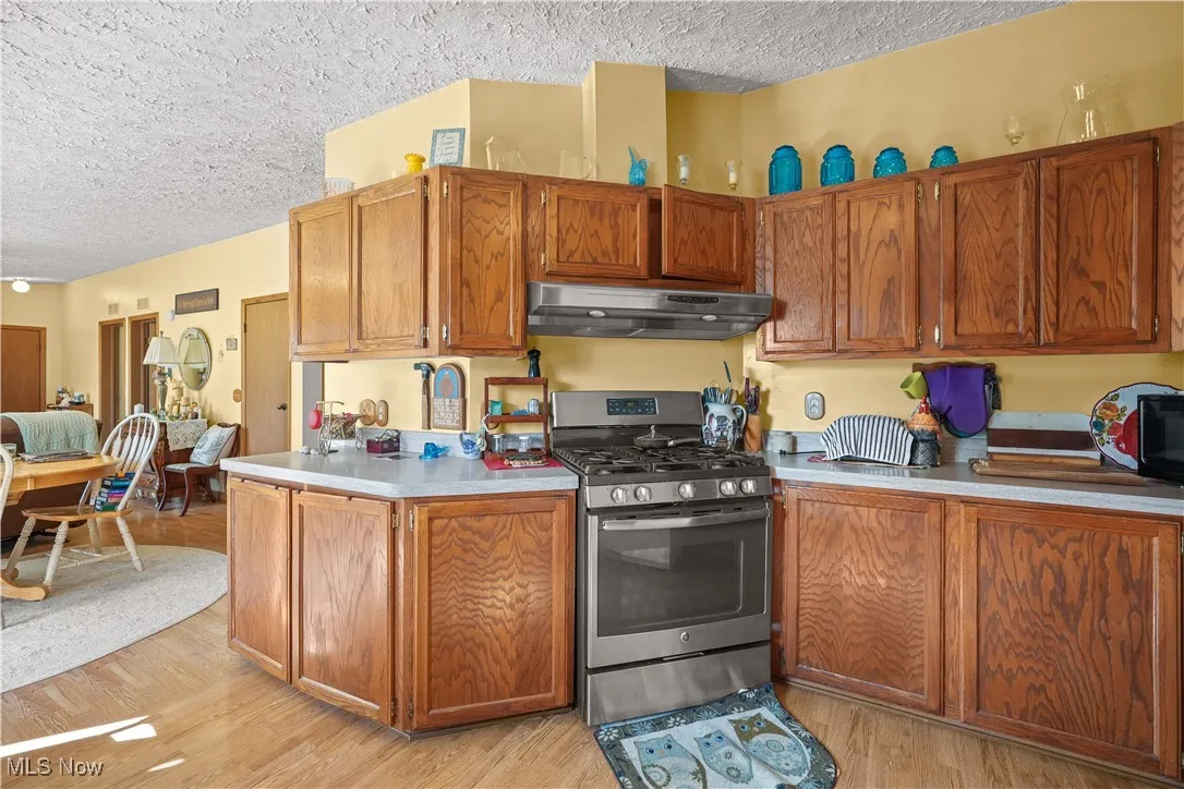 Kitchen with stainless steel gas stove, brown cabinets, a textured ceiling, light wood-type flooring, and under cabinet range hood