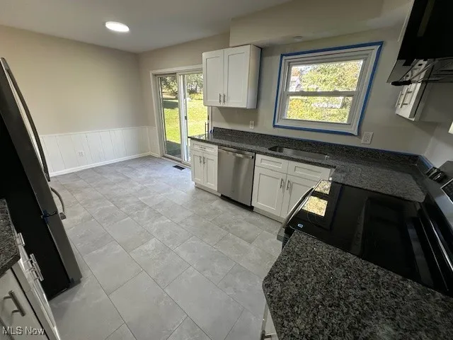 Kitchen featuring white cabinetry, dark stone counters, freestanding refrigerator, stainless steel dishwasher, and a wainscoted wall