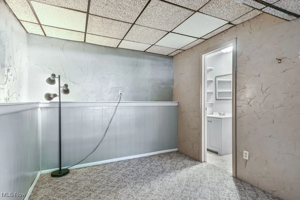 Family room with a paneled ceiling, a textured wall, light colored carpet, and a wainscoted wall. View into half bathroom.