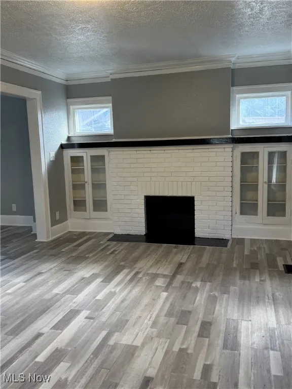 Unfurnished living room with a textured ceiling, wood finished floors, crown molding, and a brick fireplace