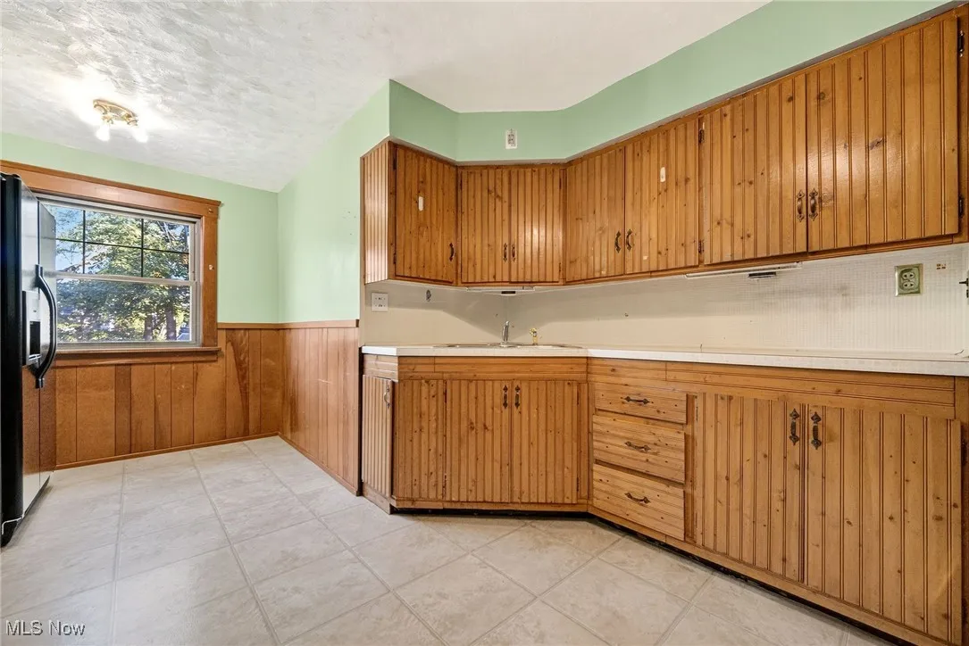 Kitchen with wainscoting, light countertops, wooden walls, black fridge, and brown cabinetry