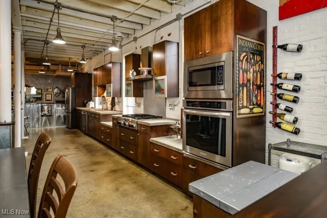 Kitchen featuring stainless steel appliances, concrete flooring, light countertops, hanging light fixtures, and wall chimney range hood