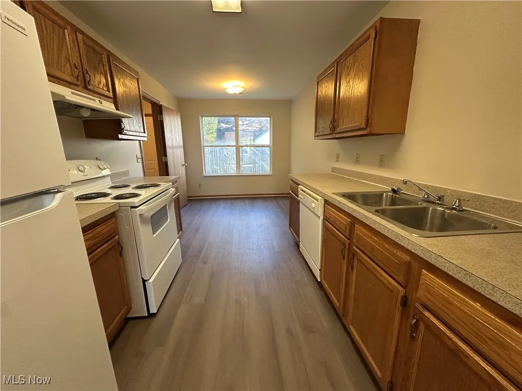 Kitchen featuring white appliances, brown cabinets, dark wood finished floors, under cabinet range hood, and light countertops