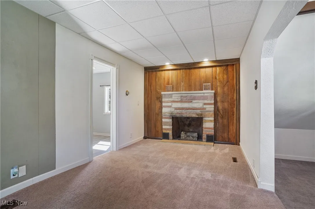 Unfurnished living room with a stone fireplace, carpet floors, wooden walls, and a drop ceiling