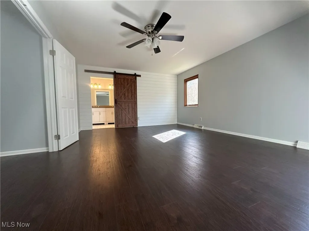 Unfurnished bedroom with a barn door, dark wood-style floors, a ceiling fan, and ensuite bathroom
