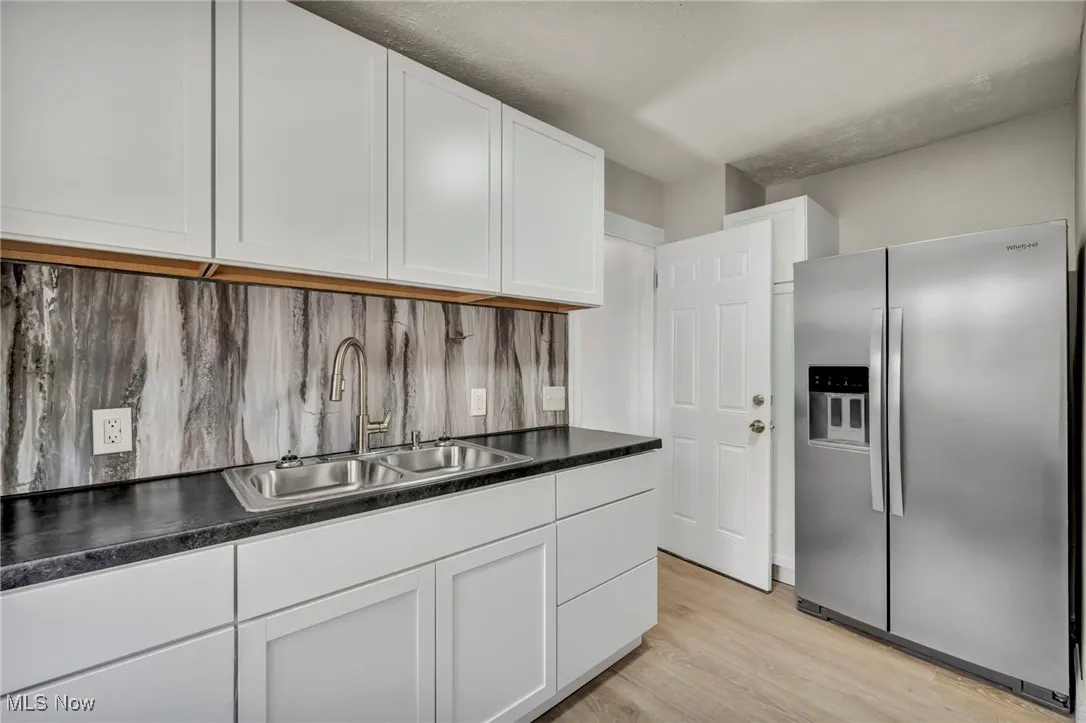 Kitchen with stainless steel fridge, light wood-type flooring, white cabinets, dark countertops, and backsplash