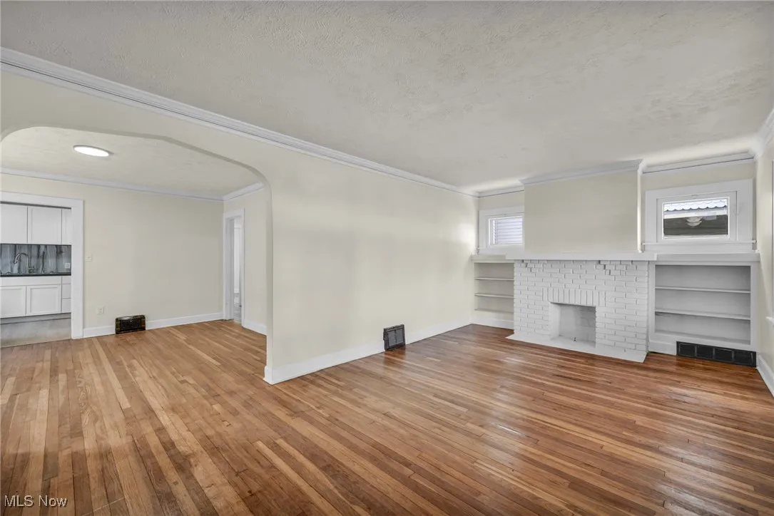 Unfurnished living room featuring arched walkways, ornamental molding, light wood finished floors, a brick fireplace, and a textured ceiling
