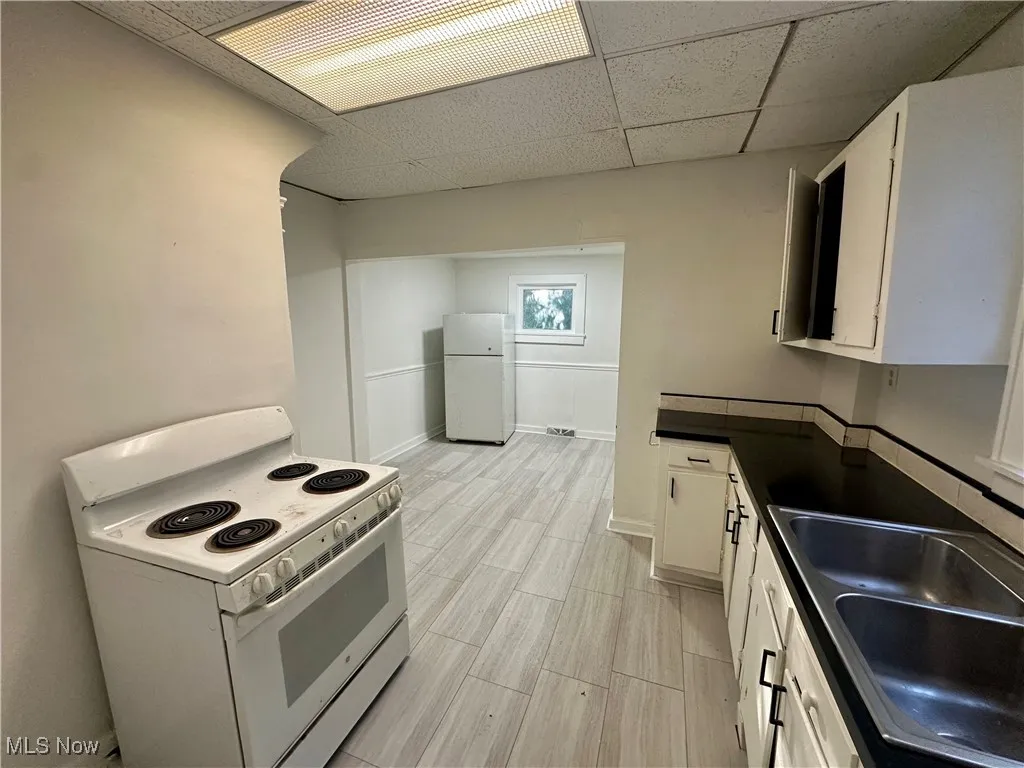 Kitchen featuring white appliances, dark countertops, a paneled ceiling, white cabinetry, and wood tiled floors