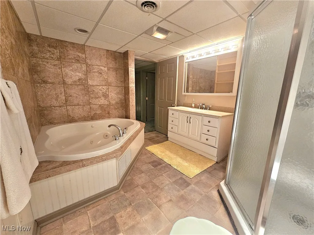 Full bath featuring a drop ceiling, a shower stall, vanity, a jetted tub, and tile walls