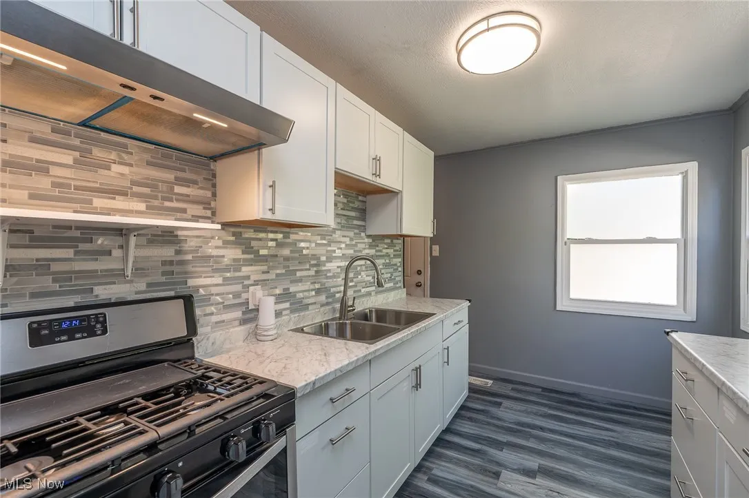 Kitchen featuring stainless steel range with gas stovetop, backsplash, under cabinet range hood, white cabinets, and dark wood-style floors
