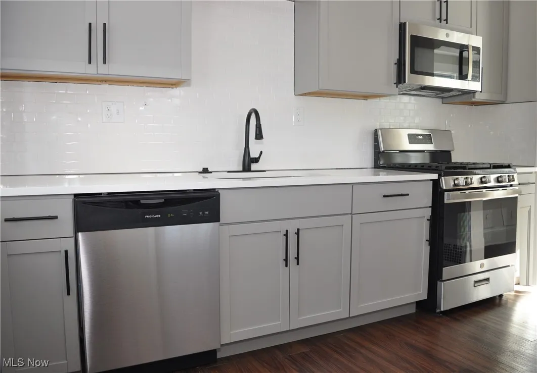 Kitchen with decorative backsplash, appliances with stainless steel finishes, gray cabinetry, and dark wood-style flooring