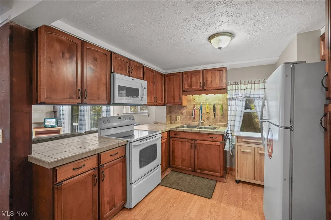 Kitchen with white appliances, light wood-style flooring, tile counters, brown cabinetry, and a textured ceiling