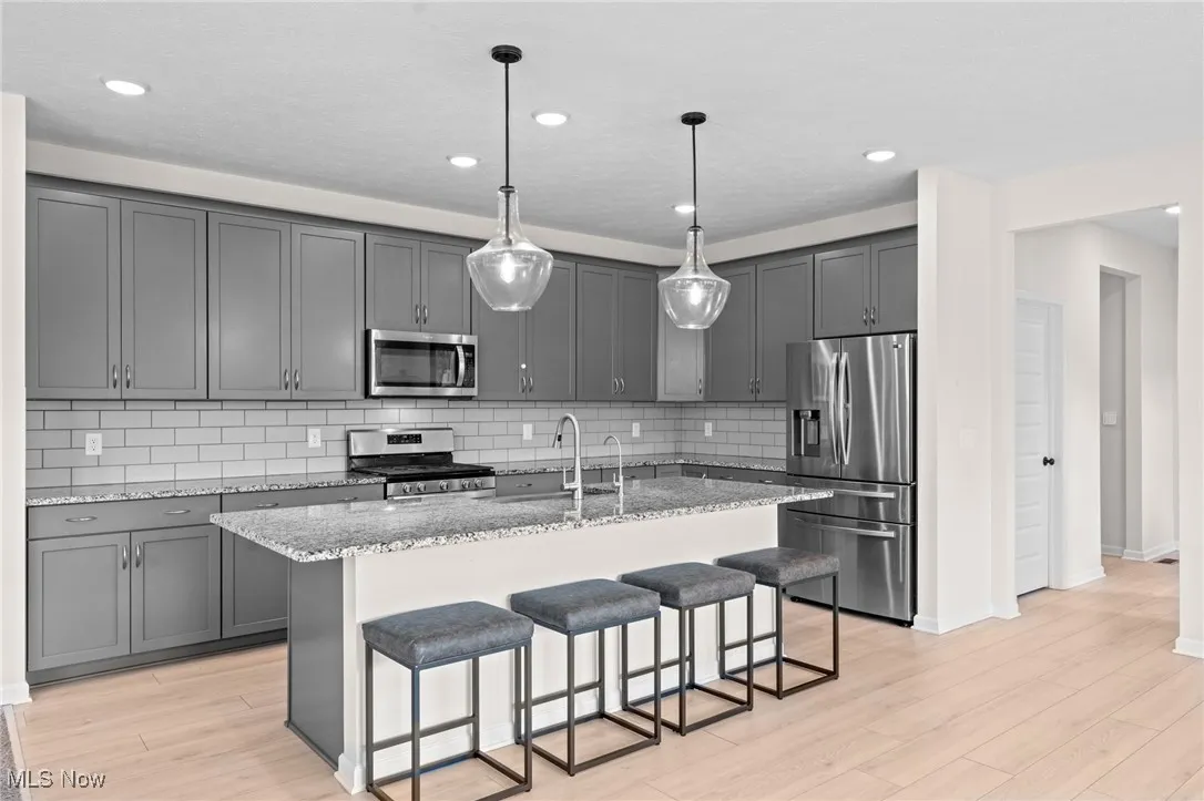 Kitchen featuring gray cabinetry, a kitchen bar, appliances with stainless steel finishes, light stone counters, and an island with sink