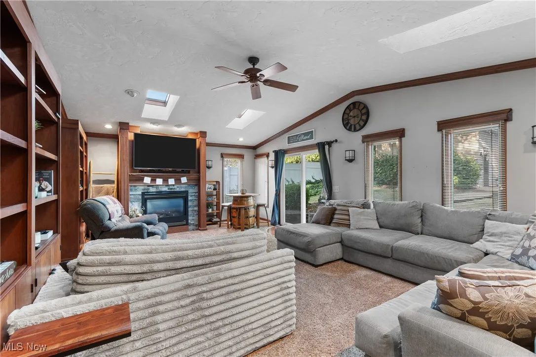 Carpeted living room featuring a skylight, ornamental molding, vaulted ceiling, a glass covered fireplace, and a ceiling fan