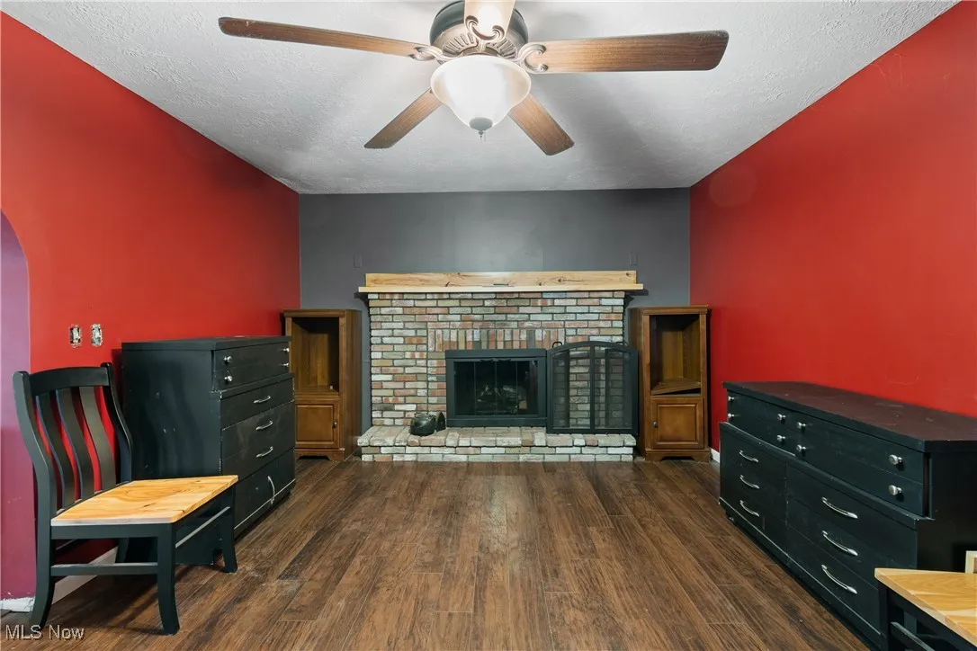 Living room with dark wood-style floors, a textured ceiling, a brick fireplace, arched walkways, and a ceiling fan