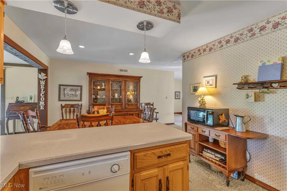 Kitchen boasting natural wood cabinetry, light colored counters, breakfast bar with pendant lighting above, window above kitchen sink overlooking backyard, white fridge, black oven, glass top electric range and light colored carpeting.