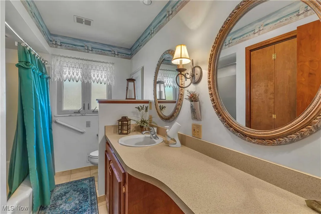 Full bathroom in the hall with a window for natural light, large updated vanity with two mirrors above, shower/bath combo with shower curtain, and light tile floors.